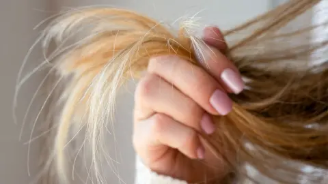Getty Images Close-up image of a woman holding a section of long blonde-brown hair, highlighting soft natural lighting and a blurred neutral background.