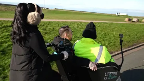 A man in a yellow jacket and a man in a black jacket sitting on a trishaw; a woman wearing a black coat, ear muffs and black sunglasses pedals behind them.