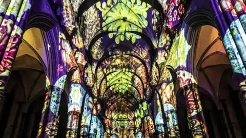 Luxmuralis installation - Lights illuminate the cathedral pillars and inside of cathedral. Purple and yellow at the front, with greens and orange and floral patterns at the back.