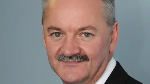 A man with grey hair and a trim moustache, looking into the camera. He is wearing a suit. 