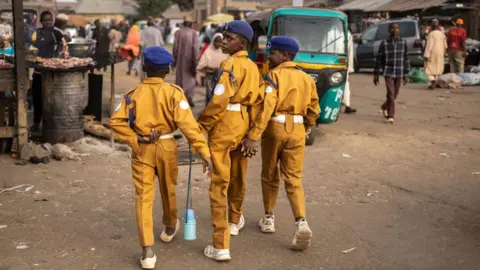 Olympia de Maismont / AFP Three boys walk away from the camera, wearing yellow trousers and shirts with a blue hats. Market traders sell goods nearby in Jos, Nigeria - Thursday 27 February 2025.