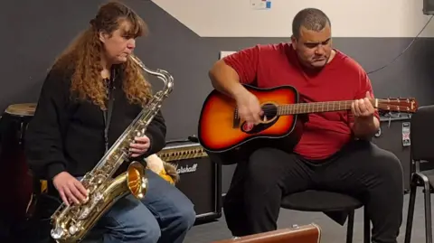 Absolute Music Trust Gaynor sits to the left, she is wearing a black long sleeved jumper and jeans, holding her saxophone as she plays music. To her right is a man with shaved black hair, wearing a short sleeved red t shirt and black tracksuit bottoms playing the guitar.