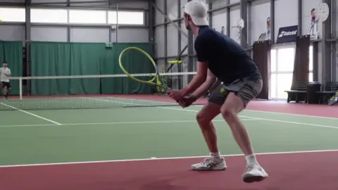 James Dunn/BBC A man with a backwards white baseball cap plays tennis at an indoor court. 