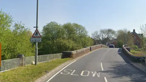 Google Google Street view of the road across Julian's Bridge. On the left is a highways sign that says 'no footway for 50 yards'. The road narrows over the hump-backed bridge.