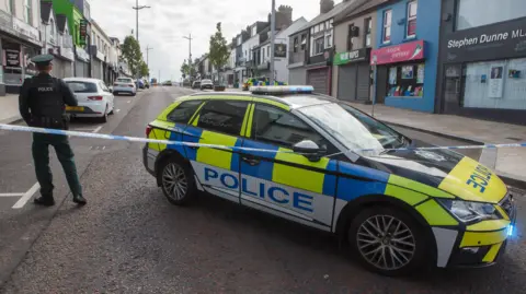 A police officer stands with their back to the camera beside a marked police car. There is a cordon in place sealing off a large main street road. There are a number of shops lining either side of the road, some with their shutters down.
