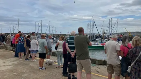 A mixed group of people wait on a harbour dock for the arrival of the Nao Victoria.