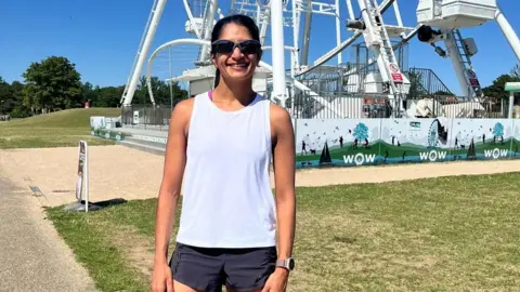 A woman stands on a sunny pathway near a large white Ferris wheel, dressed in athletic gear with bright pink running shoes.