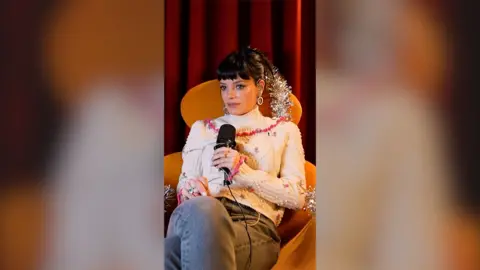 Lily Allen sits in an orange armchair with silver tinsel. She wears a white jumper with mini pink rose designs