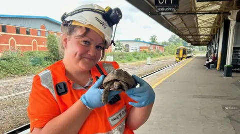 A woman wearing a helmet with a light on it, blue plastic gloves, an orange high visibility top, brown hair, smiling whilst holding a small brown tortoise between both her hands, stranding on a train platform on a cloudy day