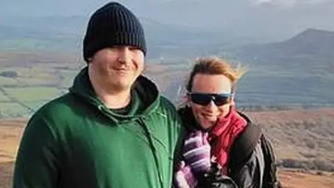Family photo Rhys and his mother, Trina Dobson, smiling and standing close together on a hilltop with the landscape of fields and hills stretched out in the distance behind them. Rhys is wearing a green hoodie and dark beanie. His mum wears a pink and black anorak, sunglassses and strippy gloves.