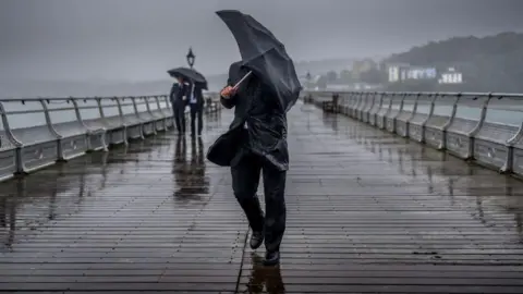 People battle against high winds and driving rain on a pier. A man is holding an umbrella which is misshaped against the wind.