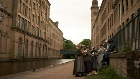 Gerontius Productions Limited A group of people in period dress gather beside a canal near a large 19th-century brick mill with tall windows and a tower.