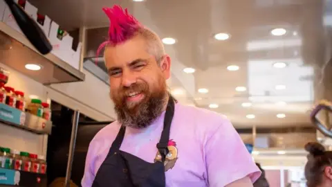 Tamworth Borough Council A man in a pink tshirt and black apron with a bright pink mohawk smiles at the camera.