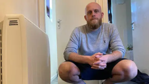 BBC Tom looks at the camera while crouching on his home's floor