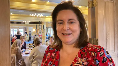 Una mujer de cabello castaño hasta los hombros sonríe con la boca abierta, de pie en una puerta de madera que da a un opulento comedor lleno de gente disfrutando del té de la tarde. Lleva un vestido rojo con flores de colores.