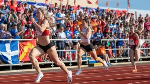 Three athletes running, about to cross the finish line. A crows of people are in the background holding various coloured island flags.