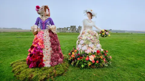 Two large sculptures depicting Victorian women in dresses made out of dahlias. They are intricate dresses with lots of different colours and textures. They are positioned on the grass a distance away from Stonehenge, which can be seen in the background on a misty day.