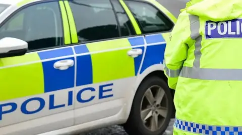 A police car and an officer with their back to the camera - we can just see the back of their hi-viz yellow jacket which has the word 'Police' on the back in white letters on a blue background. 