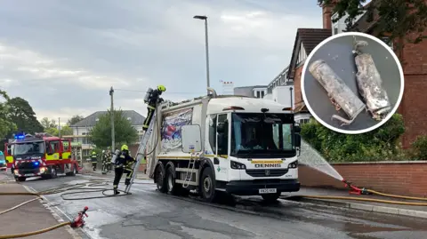 Two firefighters climb a ladder which is leaning against a bin lorry. Several hoses are also pointed at the vehicle. There is an inset picture showing a close-up of a vape which has clearly been on fire.