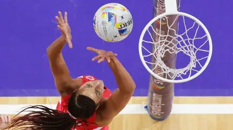 This image shows an overhead view of a netball match in progress. One player in a red kit is leaping and reaching towards a netball as it approaches the hoop.