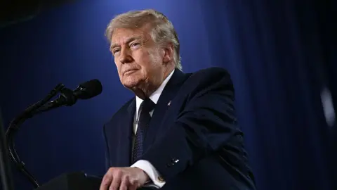President Donald Trump wears a navy suit and tie and looks off into the distance with a microphone in front of him