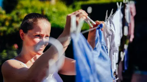 A woman wearing a white sleeveless top smiles as she hangs out a laundry on the washing line outside.