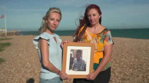 Kelly Will and Marina Polito hold a framed portrait of their brother Daniele. They are standing on a pebble beach in the sun. 