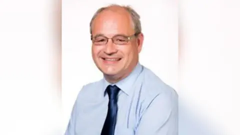 A man with grey hair and glasses smiles at the camera in front of a white background.