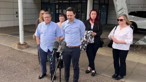 Shaun Whitmore/BBC A group of six people outside a courthouse. One man is speaking into two microphones on stands.