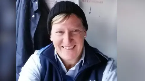A woman with blonde hair poking out from under a black beanie hat grins. She is sitting down and wearing a blue shirt and navy bodywarmer.