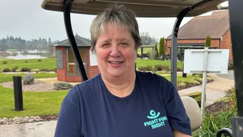 A woman sitting in a golf buggy. She is wearing a navy t-shirt that has a turquoise logo that reads "Fight for Sight". She has short hair with blonde and dark brown highlights. She is smiling and a golf course with a lake can be seen in the background. 
