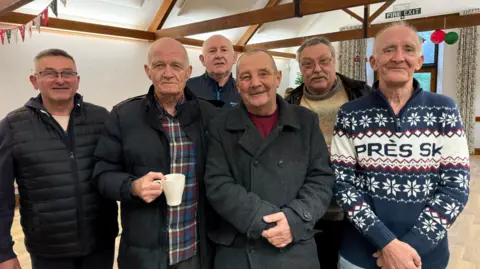 Six male veterans stand in line with arms crossed and smile at the camera.