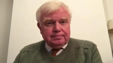 BBC A picture of Dr Tim Brain - a screenshot of an interview. He is wearing a shirt, red tie and a green jumper on top.