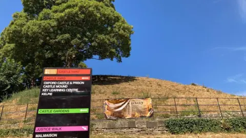 A large sign to the front left of the photo helps direct tourists to various parts of Castle Quarter. Behind it is a large mound with a tree on top.