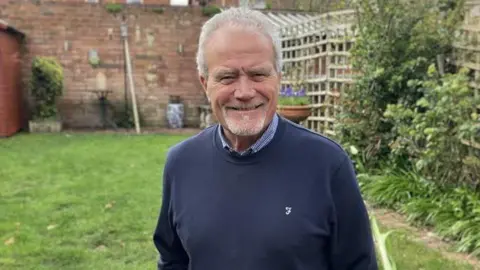 Mark Ansell/BBC Brunt in a well‑kept garden. He is wearing a dark blue sweater layered over a collared shirt and has both hands in the pockets. He is positioned on a paved or stone surface that borders a grassy lawn.