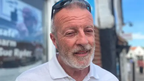 A man with a grey beard and sunglasses on top of his head squints into the camera on a bright day.