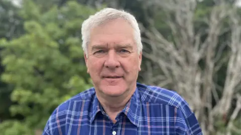 Portrait of a person with short grey hair wearing a blue check-pattern shirt, standing outdoors in a grassy garden area. Trees and shrubs form a blurred background, with a leafless tree visible to the right, and a mobile phone partially visible in the shirt pocket.