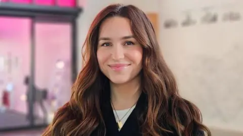 Amy stands in a building entrance hall and smiles at the camera. She has long brown hair that is slightly curled. She wears a black coat with a white top underneath.