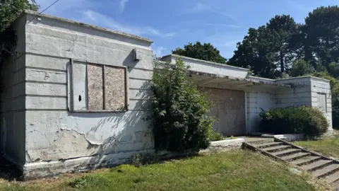 An image of the entrance to the Broomhill Lido. It appears in a debilitated state with large bushes growing at the front of it and the doorway boarded up. 