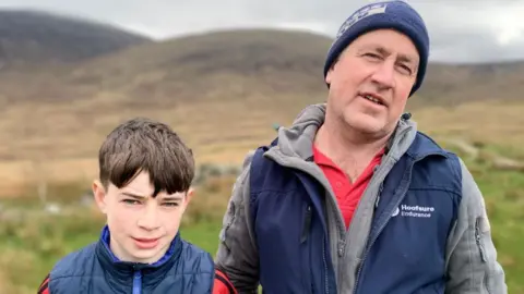 A young boy with brown hair and wearing a red t-shirt with a navy gilet over is is looking at the camera. He is on the left of the image. On the right is a man with a blue woolly hat on his head. He's wearing a red t-shirt, grey hoodie and navy gilet.