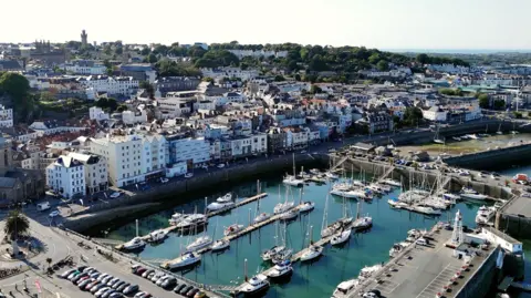 The picture shows a coastal town with a busy harbour filled with yachts and small boats. The water is a blue-green, and the marina is surrounded by stone harbour walls and long rows of parked cars. Beyond the harbour there is a tightly packed cluster of buildings, most of them light-coloured and a few storeys high, rising up the hillside. The town stretches inland towards greener areas with trees and open spaces further back. 