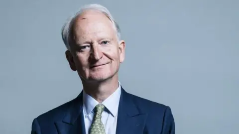 Chris McAndrew Sir Henry Bellingham has white hair and a mark on his upper forehead. He is smiling. He is wearing a blue jacket, light blue shirt and a green patterned tie. There is a handkerchief in his breast pocket. The background is plain light blue.