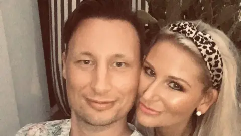 Mark Beesley A man with short brown hair and a white floral shirt takes a selfie of him and a woman with long blonde hair and a black and white headband.