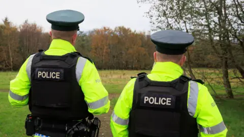 Two PSNI officers have their backs to the camera. They have on yellow police coats and black body armour that says 'police' on the back.