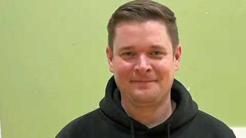 Benjamin Gillham has short brown hair and is wearing a black hooded top with a school logo and the word staff written on it. He is stood in a room with a green painted wall and microphone