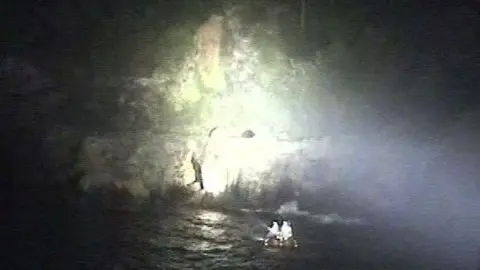 Torbay RNLI An RNLI crew inspects a stretch of coastline during a rescue operation. A spotlight is shining on part of the coastline.