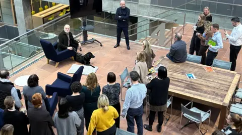 BBC News staff gather in the Glasgow studios to say goodbye to Major. The dog is lying in front of Ian Hamilton, who is sitting on a navy blue armchair next to Howard Simpson, head of news who is standing next to a TV screen.