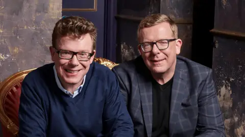 Twin brothers Craig and Charlie Reid, both wearing glasses and smiling