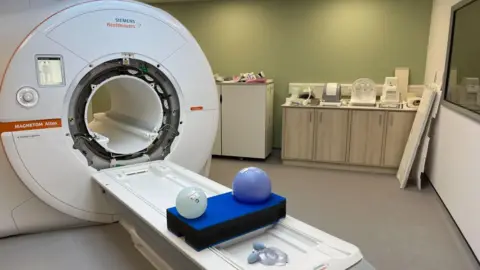 University Hospitals of North Midlands NHS Trust General view of an MRI maching inside a hospital room with grey floors and olive green wall.