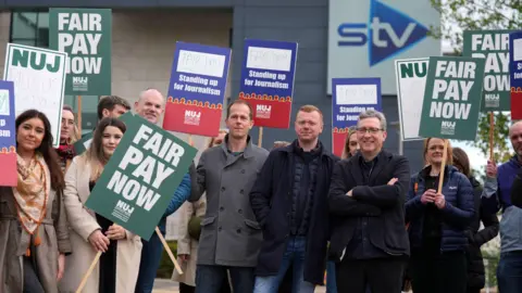  A group of people holding placards outside a building with the STV logo. Signs read “Fair Pay Now” and “Standing up for Journalism,” representing a protest organized by the National Union of Journalists (NUJ).